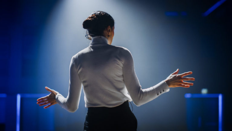 public speaking tips | woman in white turtleneck speaking to an audience, back to the camera