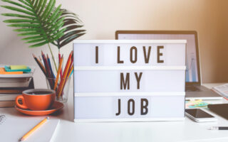 psychological safety | lightbox sign that reads "I Love My Job" on top of a desk with a computer, jar of pencils and a cup of coffee