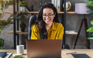 email etiquette | woman in yellow shirt sitting at desk with laptop, there are plants behind her on a bookshelf