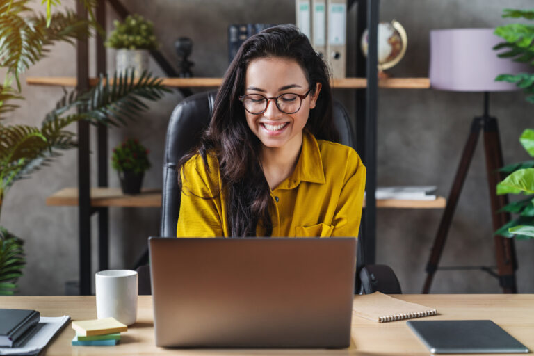 email etiquette | woman in yellow shirt sitting at desk with laptop, there are plants behind her on a bookshelf