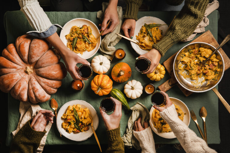 Friendsgiving Etiquette | Table overhead with pumpkins and plates of Thanksgiving food, friends making a toast