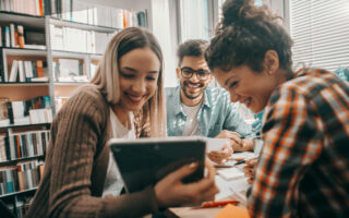 social media etiquette refresh | Three happy students dressed casual using tablet for school project and sitting at desk in library