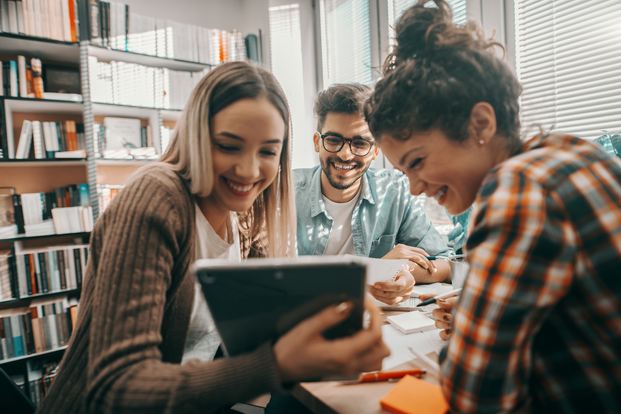 social media etiquette refresh | Three happy students dressed casual using tablet for school project and sitting at desk in library