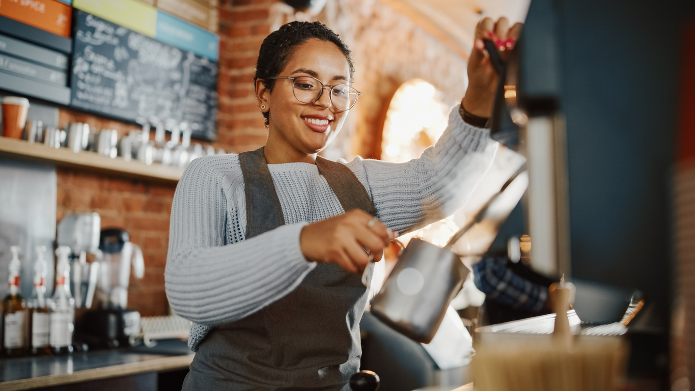 employee happiness | Latin American Female Barista with Short Hair and Glasses is Making a Cup of Tasty Cappuccino in Coffee Shop Bar. Male Cashier Works at a Cozy Loft-Style Cafe Counter in the Background