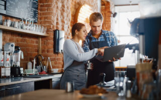 employee happiness | Two Diverse Entrepreneurs Have a Team Meeting in Their Stylish Coffee Shop. Barista and Cafe Owner Discuss Work Schedule and Menu on Laptop Computer. Multiethnic Female and Male Restaurant Employees.