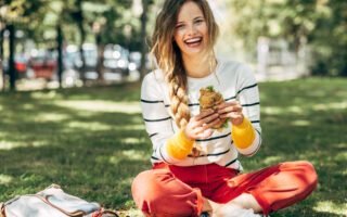 Why You Should Avoid Eating Lunch At Your Desk | Happy student female sitting on the green grass at the college campus on a sunny day, have lunch and studying outdoors. A smiling young woman takes a rest eating fast food and learning in the park.