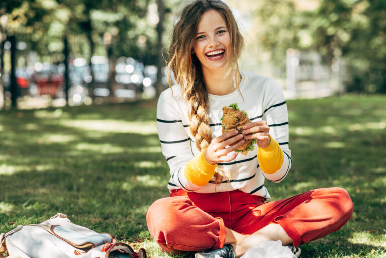 Why You Should Avoid Eating Lunch At Your Desk | Happy student female sitting on the green grass at the college campus on a sunny day, have lunch and studying outdoors. A smiling young woman takes a rest eating fast food and learning in the park.