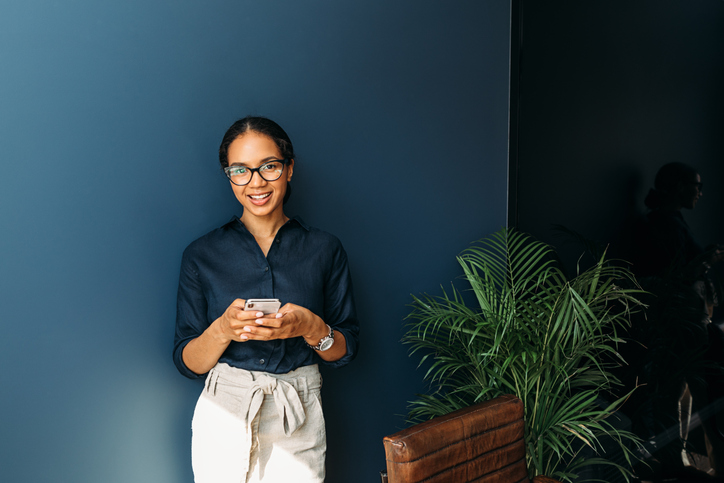 responding to texts | technology etiquette | Beautiful smiling woman looking at camera while using a smartphone at a home office