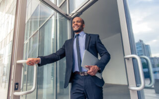 How to Look for a Job | Low angle view of Black businessman in classic suit holding a laptop and smiling while leaving the office building
