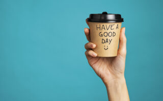 positive mindset | Close up of hand of young woman holding take away coffee cup with text written on it. Woman hand holding a coffee paper cup isolated on blue background. Morning disposable mug with message of good day written over it.