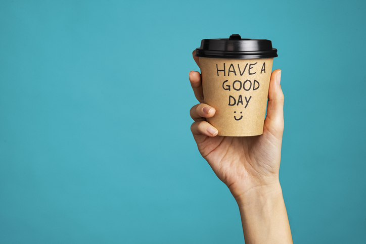 positive mindset | Close up of hand of young woman holding take away coffee cup with text written on it. Woman hand holding a coffee paper cup isolated on blue background. Morning disposable mug with message of good day written over it.