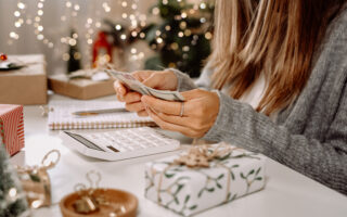 2022 Holiday Tipping Guide | woman counting US Dollar bills, using calculator, and writing expenses. Woman doing budget, estimating money balance for holiday shopping. Woman counting Christmas gifts.