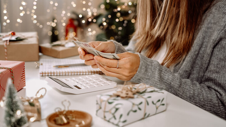 2022 Holiday Tipping Guide | woman counting US Dollar bills, using calculator, and writing expenses. Woman doing budget, estimating money balance for holiday shopping. Woman counting Christmas gifts.