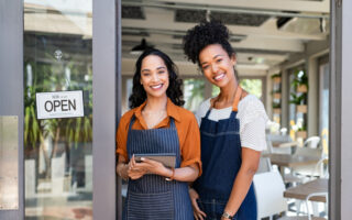 Portrait of smiling young latin woman holding digital tablet with black colleague at cafe entrance door. Two small business owners standing together at cafe entrance while smiling. Happy successful multiethnic small business women wearing apron and standing with open sign at entrance gate of restaurant.