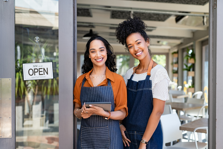 New Year's Resolutions for Entrepreneurs | Portrait of smiling young latin woman holding digital tablet with black colleague at cafe entrance door. Two small business owners standing together at cafe entrance while smiling. Happy successful multiethnic small business women wearing apron and standing with open sign at entrance gate of restaurant.