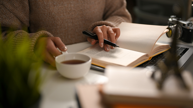 New Year's Resolutions Check-In | A female reading a book or writing a diary and sipping hot tea in cozy home workspace. leisure and hobby concept