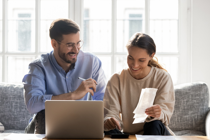 Head shot happy young family couple planning monthly budget, sitting together on cozy sofa, using computer software. Smiling mixed race wife calculating income and outcome together with husband.