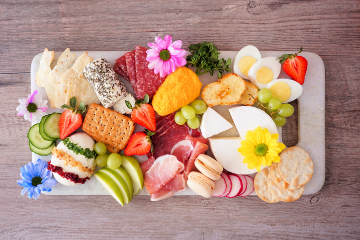 Spring or Easter theme charcuterie board against a wood background. Variety of cheese, meat, fruit and vegetable appetizers. Overhead view.