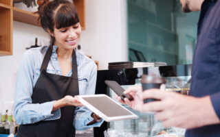 2023 Tipping Guide | Young barista woman is holding tablet for customer using smart phone scan QR code on tablet for payment at counter bar at coffee shop. Technology of digital pay without money concept.