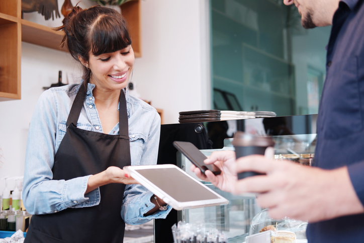 2023 Tipping Guide | Young barista woman is holding tablet for customer using smart phone scan QR code on tablet for payment at counter bar at coffee shop. Technology of digital pay without money concept.