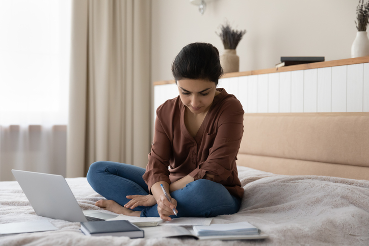 Establish a budget | Concentrated young woman sitting on cozy bed, involved in studying distantly on computer, writing notes in copybook, preparing for professional test examination, improving knowledge remotely.