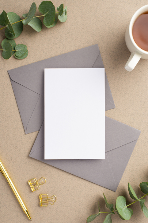 Workspace concept. Top view vertical photo of paper sheet two grey envelopes cup of tea gold pen binder clips and eucalyptus on beige background with copyspace