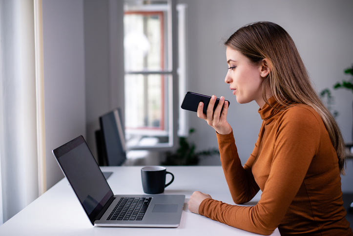 Young woman holding cell phone and recording her voice while working at the desk in the office. Leaving voice message.