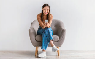 The New Cell Phone Etiquette | Happy Caucasian woman using cellphone, chatting on internet, working or studying online, sitting in armchair against white studio wall, full length. Cheery young lady watching video on mobile phone