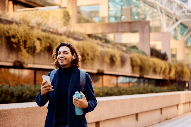 Happy Hispanic businessman using earbuds and cell phone walking outdoors.