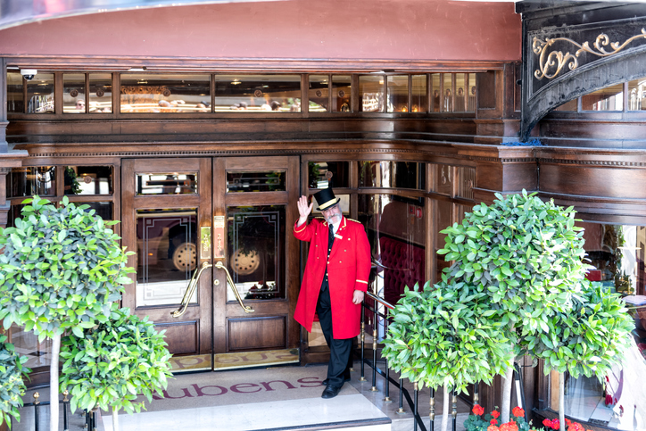 London, UK - June 22, 2018: Porter, doorman in traditional attire, clothing, hat, red gown, coat standing at Rubens hotel, inn entrance waving hand