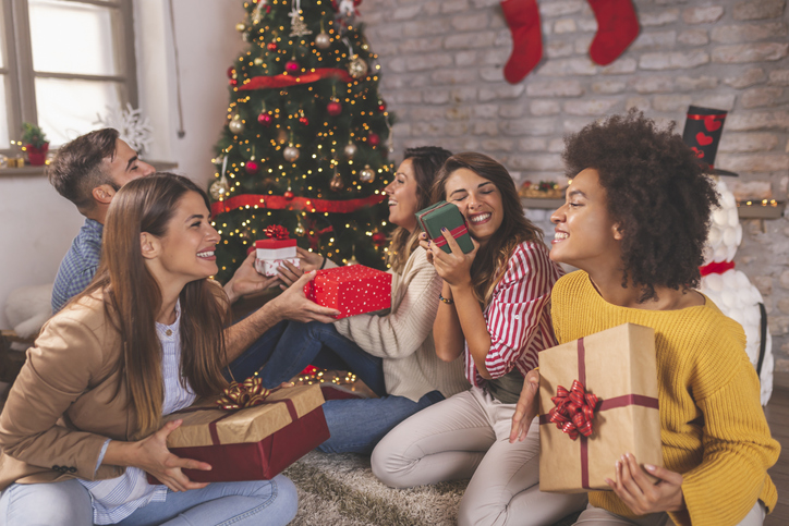 Group of friends having fun while celebrating Christmas at home, sitting by nicely decorated Christmas tree and exchanging gifts