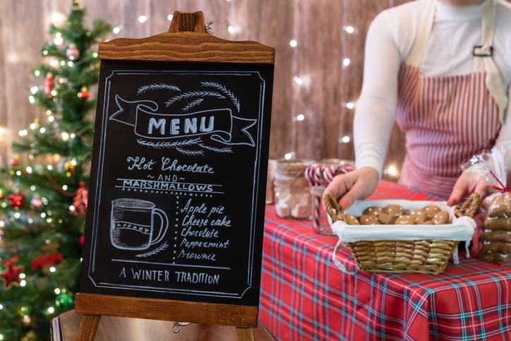 Christmas pastry shop . Woman seller, waitress selling gingerbread, marshmallows, cookies, sweets and hot chocolate in a small cozy cafe. Homemade bakery menu in the foreground