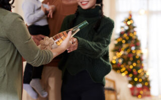 Holiday Houseguest Etiquette 2023 | Hands of senior woman giving decorated bottle of champagne or wine to adult daughter as Christmas present