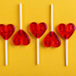 Spark Joy This Valentine’s Day | Five red sweet tasty lollipops in shape of heart on bright yellow background. St. Valentine's day.