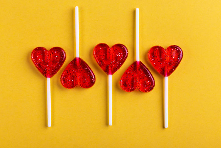 Spark Joy This Valentine’s Day | Five red sweet tasty lollipops in shape of heart on bright yellow background. St. Valentine's day. 