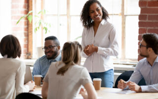 Is workplace etiquette on the decline? Multinational businesspeople gathered together at modern office conference boardroom partners during business meeting, black african female team leader ready to speak standing looking at colleagues