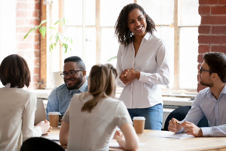 Is workplace etiquette on the decline? Multinational businesspeople gathered together at modern office conference boardroom partners during business meeting, black african female team leader ready to speak standing looking at colleagues