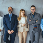 Fall 2024 Internship Q&A | The business people standing on the gray wall background. Business team headed with boss, posing to camera over grey wall in office. Diverse businesspeople smiling at the camera