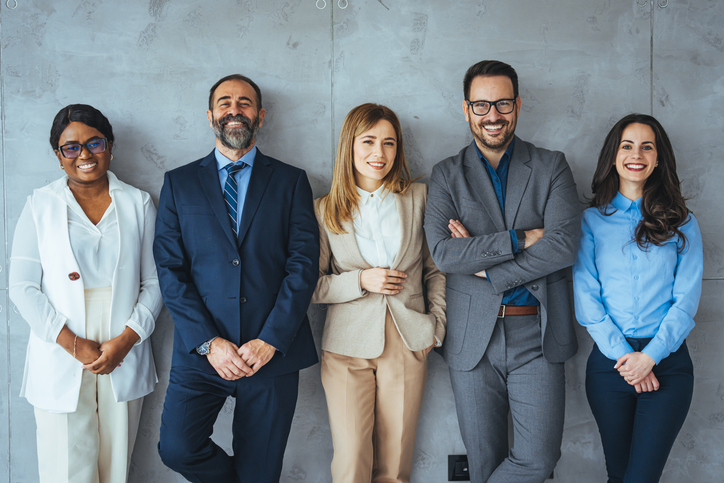 Fall 2024 Internship Etiquette Q&A | The business people standing on the gray wall background. Business team headed with boss, posing to camera over grey wall in office. Diverse businesspeople smiling at the camera