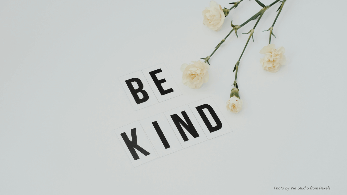 Compassion in Crisis | Black block letters that say "Be Kind" next to pink flowers, mostly white background