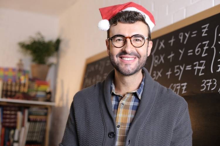 Teacher wearing santa hat and sweater smiling, standing in front of chalkboard