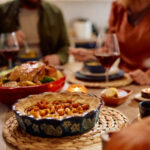 Thanksgiving Potluck Etiquette | Traditional pumpkin pie with couple eating Thanksgiving dinner in the background