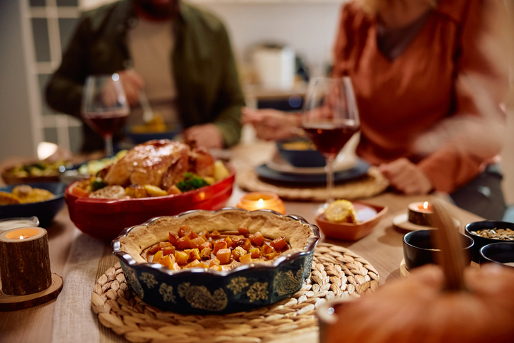 Thanksgiving Potluck Etiquette | Traditional pumpkin pie with couple eating Thanksgiving dinner in the background
