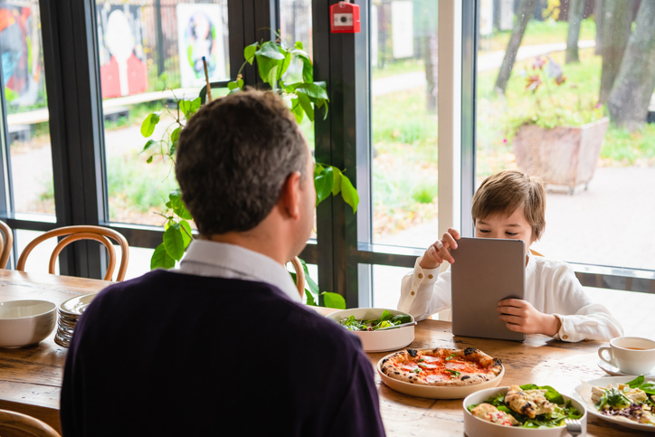 A boy sitting at the kitchen table in front of his father with a tablet.