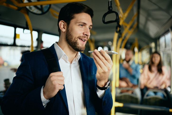 Happy businessman recording voice message on smart phone while commuting to work by bus.