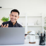 Video interview etiquette | Smiling businessman holding a notebook while having a video call meeting on his laptop in a modern office.