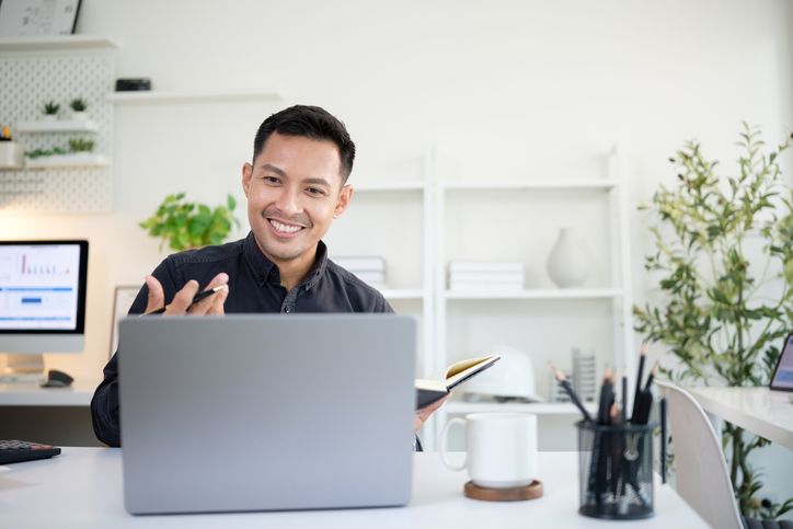 Video interview etiquette | Smiling businessman holding a notebook while having a video call meeting on his laptop in a modern office.