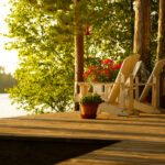 Lake house entertaining etiquette | White Adirondack chairs sits on a cottage wooden deck at sunset looking at the lake. Flower pots are on the deck.