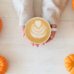 Pumpkin Spice Fall Etiquette Questions Pumpkin latte in a hands of woman | Cropped shot of young woman in a sweater holding a cup of latte with foam art. Female with a hot coffee drink and traditional autumnal holidays decoration. Close up, copy space, top view, background.