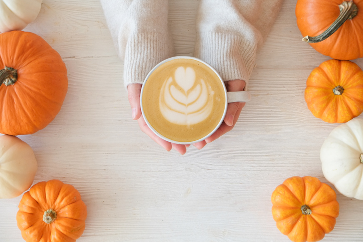 Pumpkin Spice Fall Etiquette Questions Pumpkin latte in a hands of woman | Cropped shot of young woman in a sweater holding a cup of latte with foam art. Female with a hot coffee drink and traditional autumnal holidays decoration. Close up, copy space, top view, background.